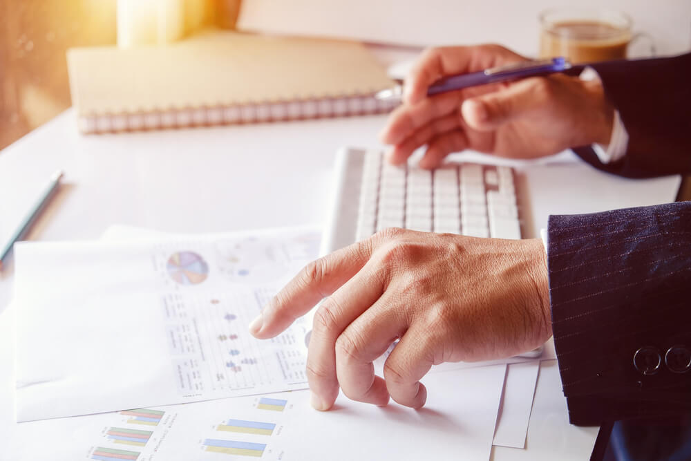 A person in a suit uses a keyboard while analyzing printed charts and graphs on a desk.