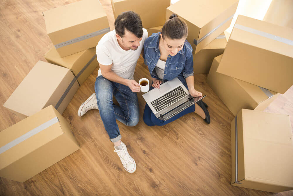 A couple sits on the floor surrounded by moving boxes, using a laptop and enjoying a cup of coffee.