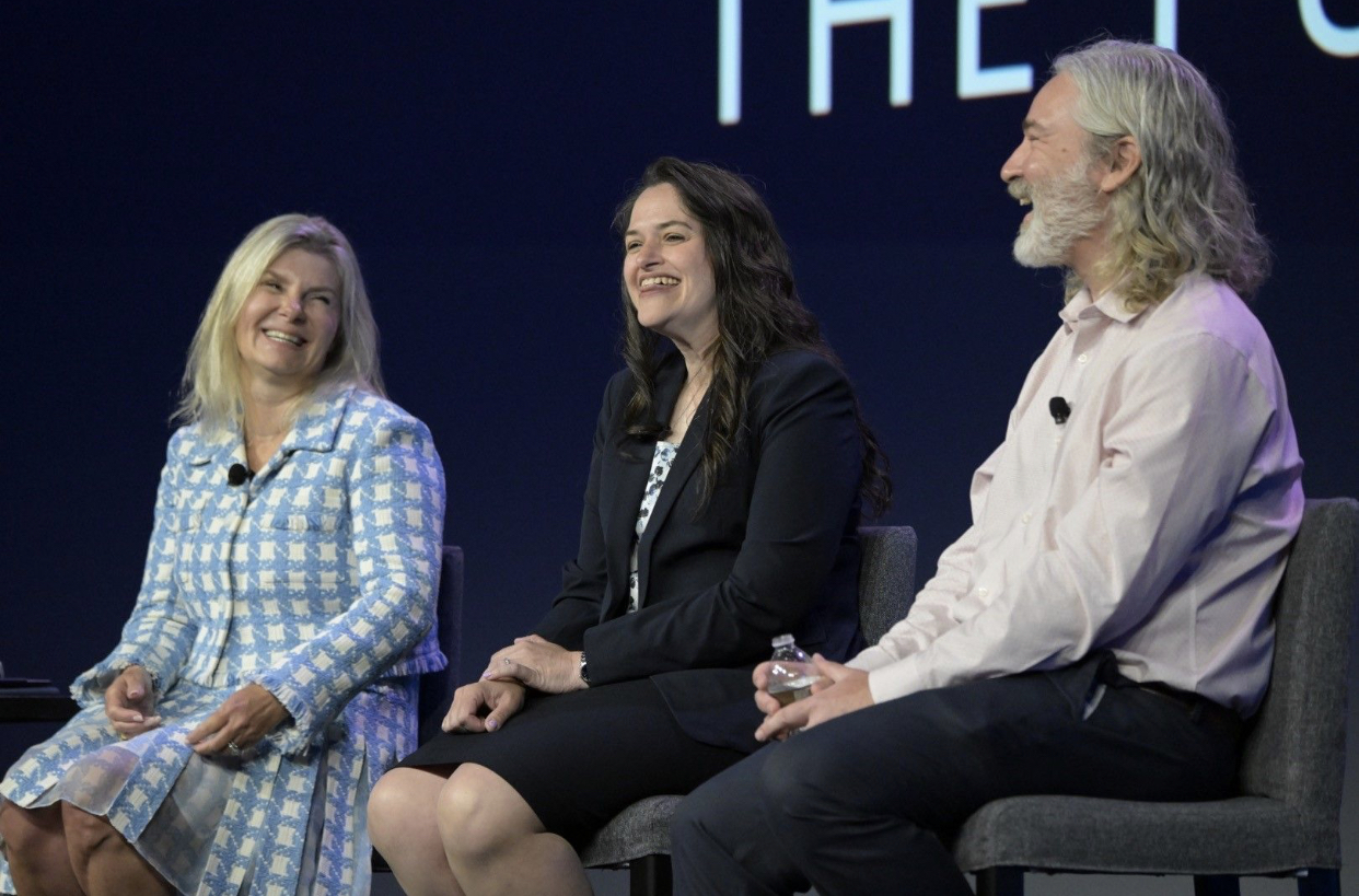 Three speakers on a panel, smiling and engaged in discussion, with a dark background and large text behind them.