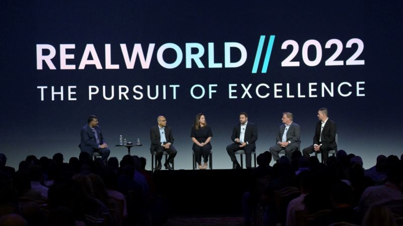 A panel discussion at RealWorld 2022 featuring five speakers on stage, with a backdrop displaying the event's title.