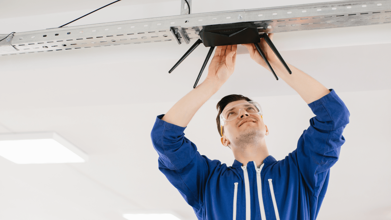 A man installs a black router onto a metal bracket on the ceiling.