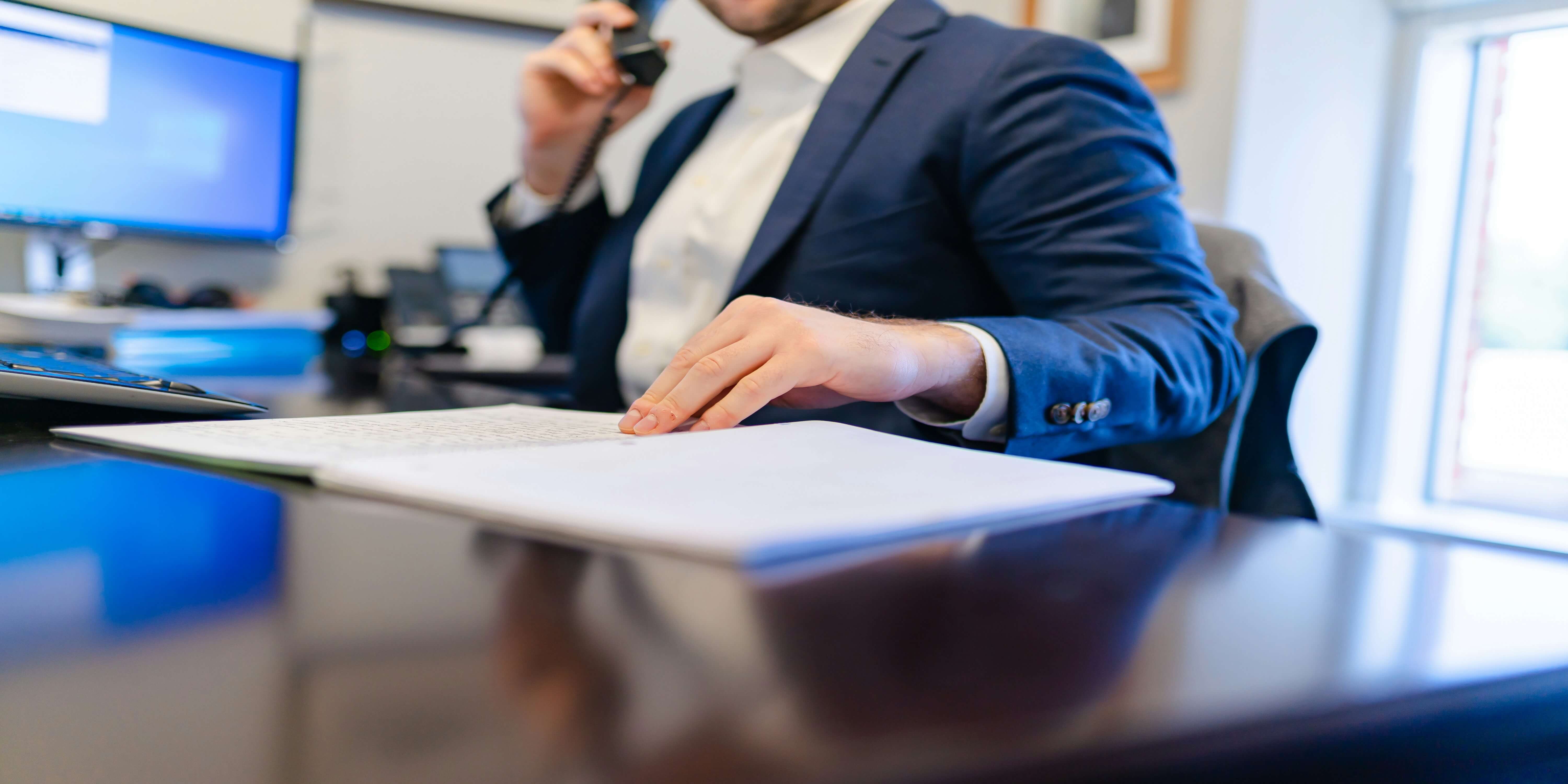 A person in a suit at a desk, on a phone, with notes and a computer monitor in the background.
