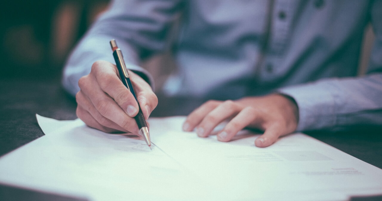 A person writing on a document with a pen, their hand visible on a table.