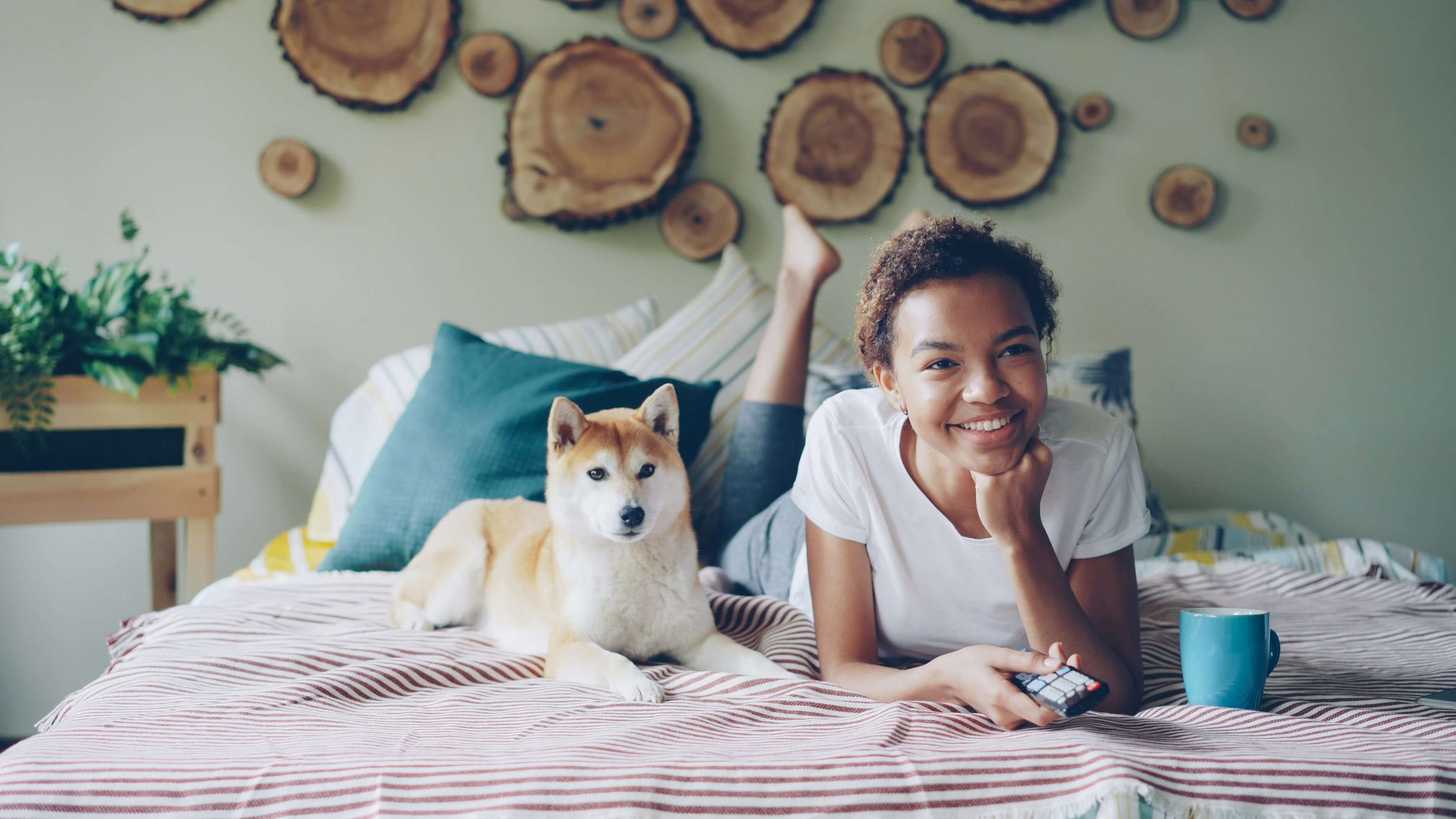 A young woman smiles while lying on a bed next to a Shiba Inu dog, with decorative wooden wall art in the background.