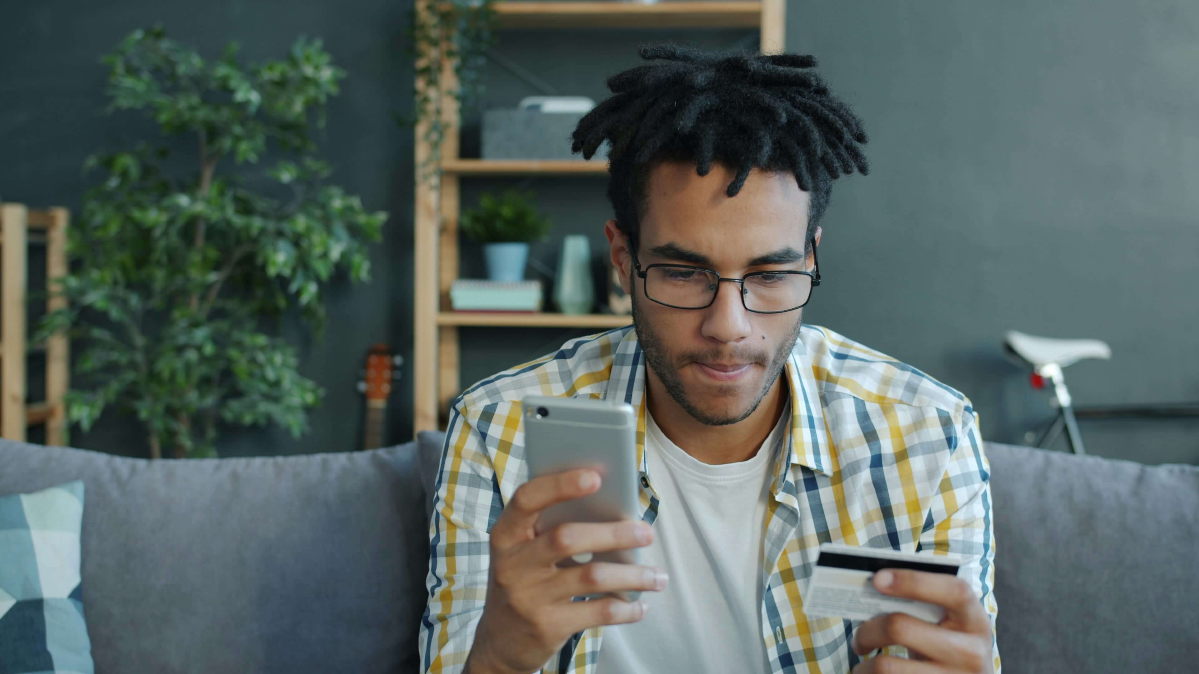 A young man with glasses holds a smartphone and a credit card while sitting on a couch, focused on a task.