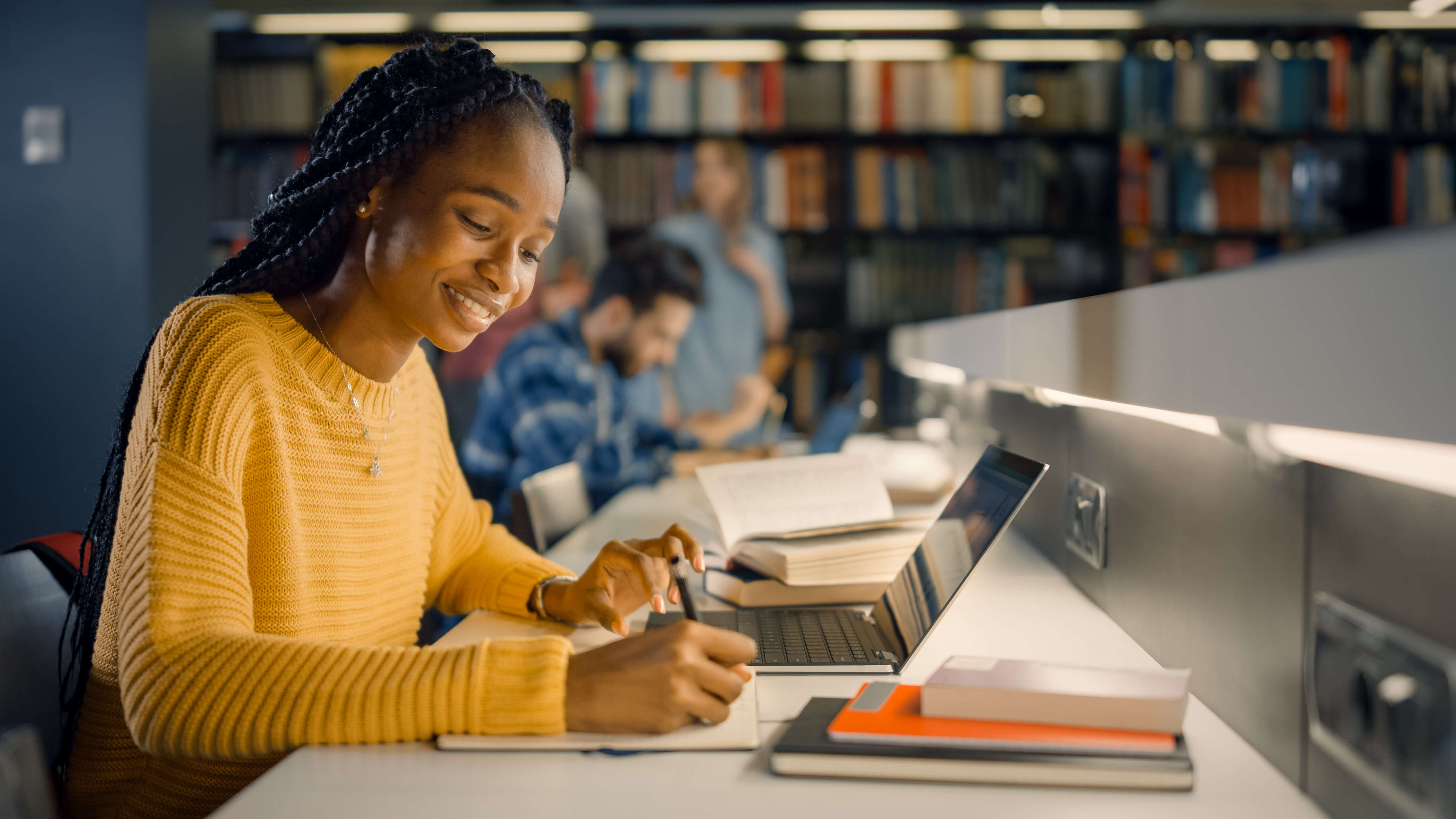 A smiling student in a yellow sweater writes notes while studying at a desk in a library.