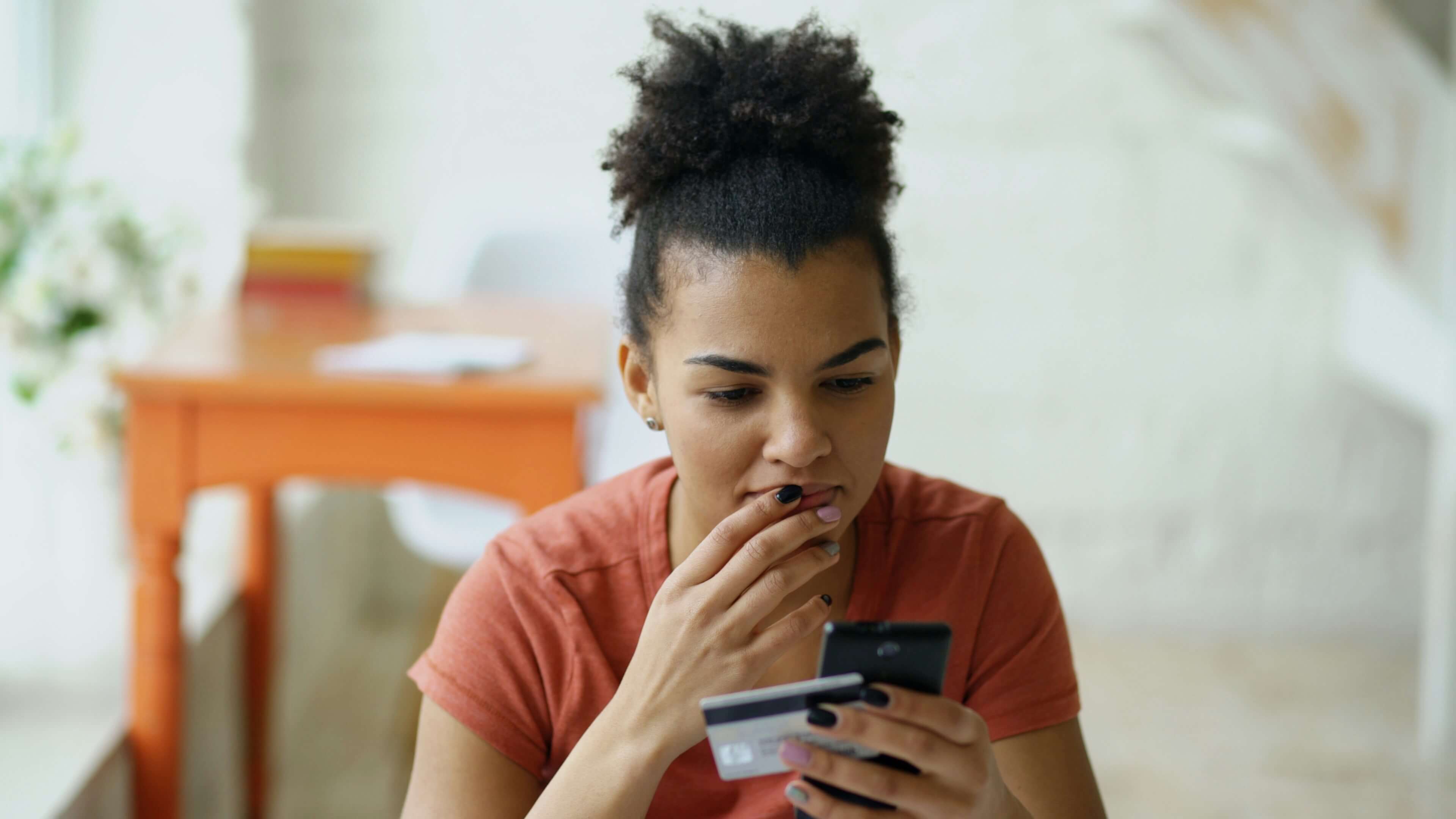 A young woman with curly hair looks at her mobile phone while holding a credit card, deep in thought.