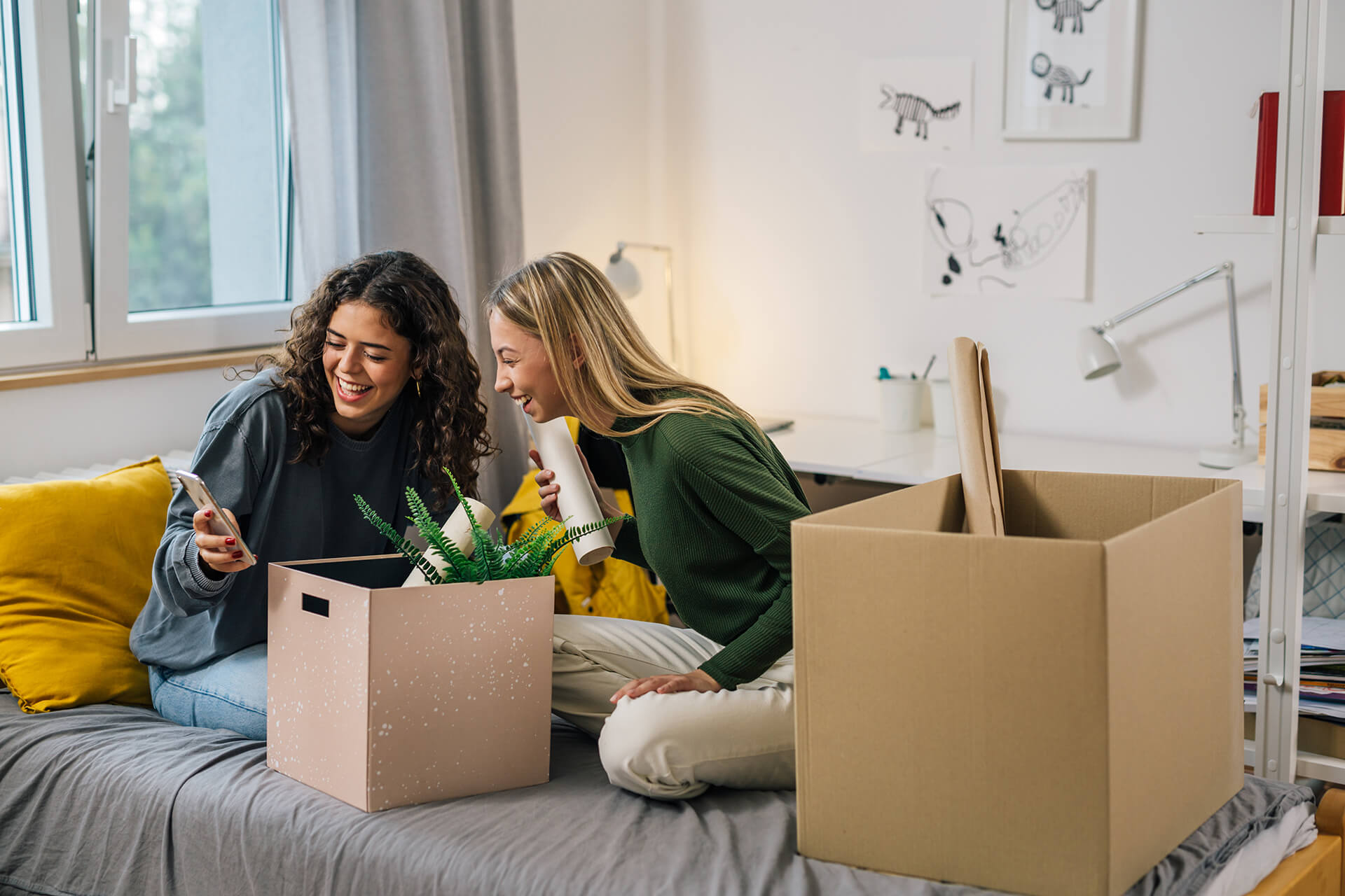 Two women sitting on a bed, smiling and looking at a phone, with moving boxes and a plant nearby.