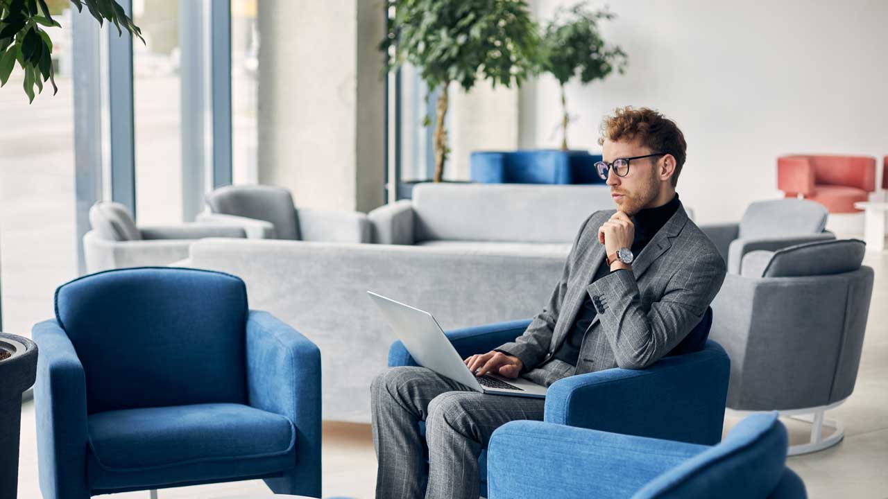 A business professional in a gray suit sits thoughtfully in a blue chair, working on a laptop.