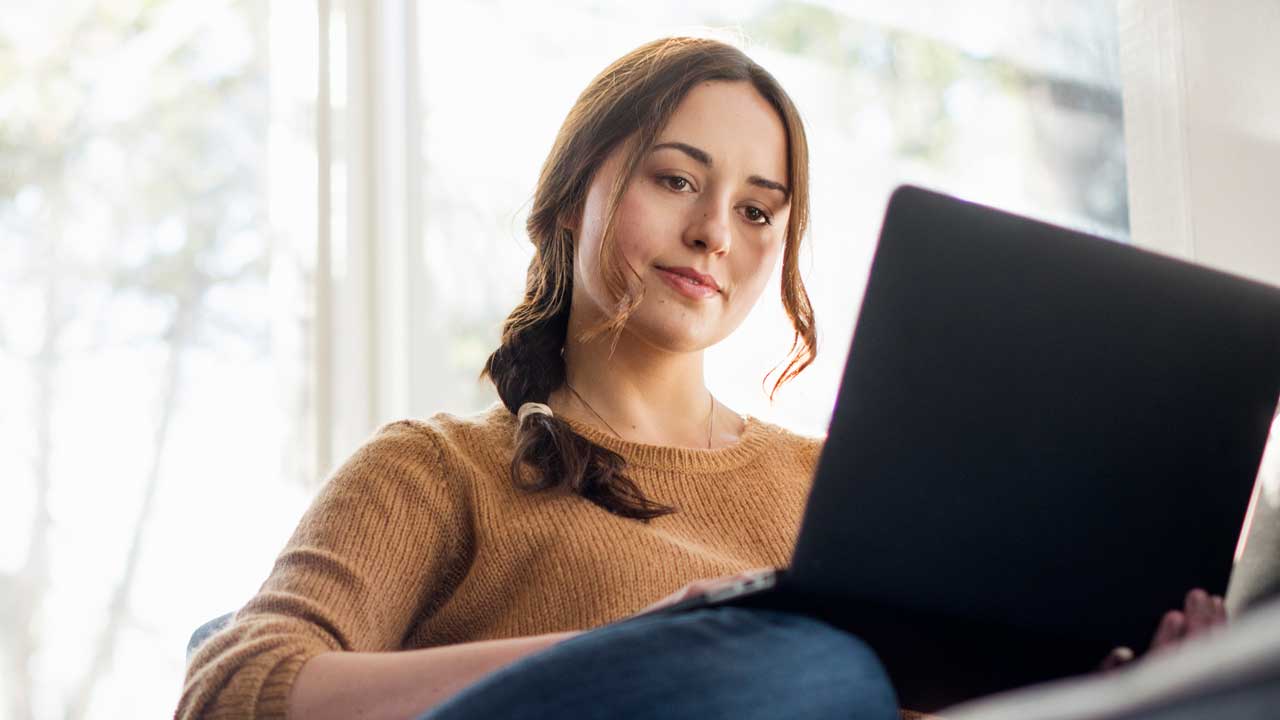 A woman with long hair is sitting comfortably while using a laptop in a bright, airy room.