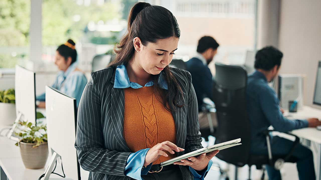 A woman in a pinstriped suit and orange sweater works on a tablet in a modern office with colleagues in the background.