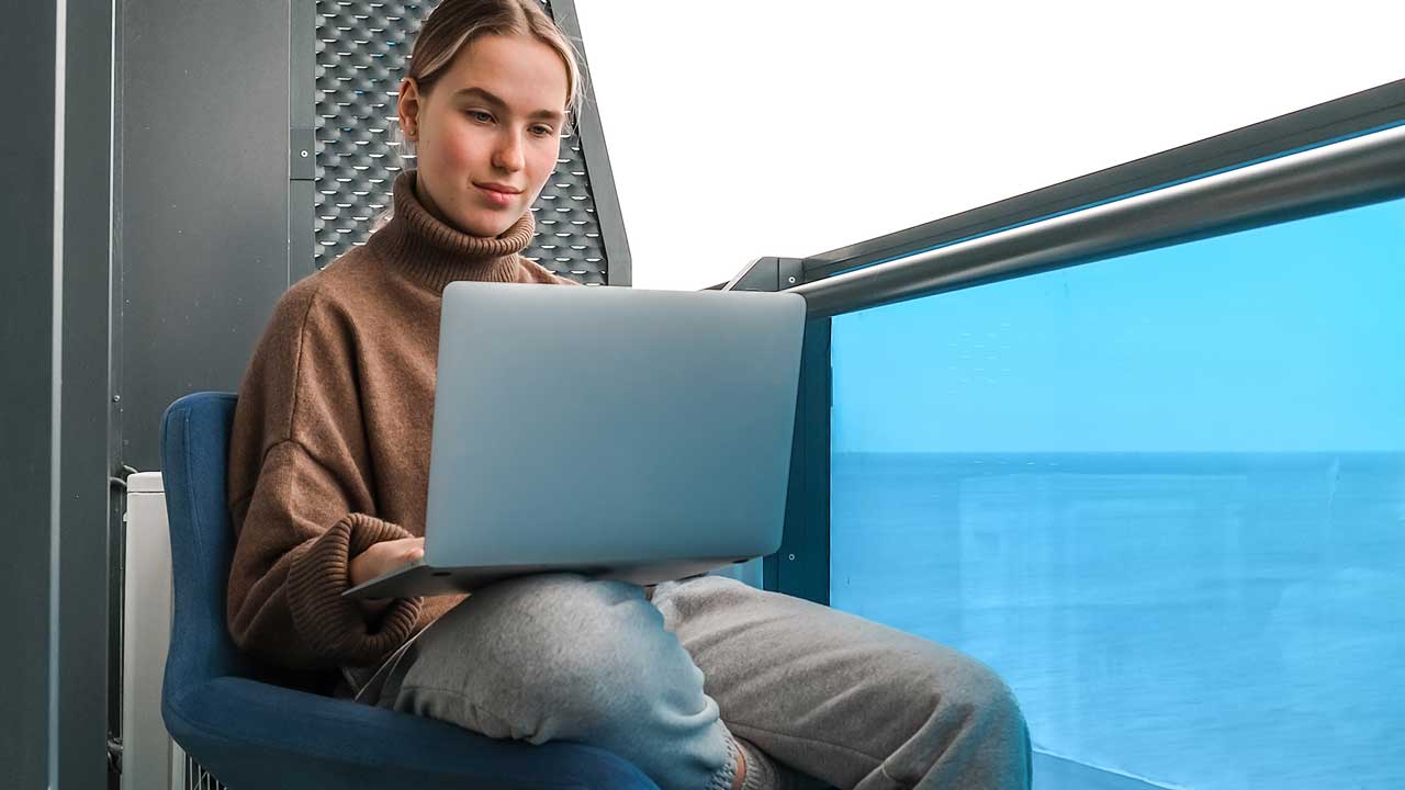 A young woman in a brown sweater uses a laptop while sitting on a balcony with a view of the ocean.