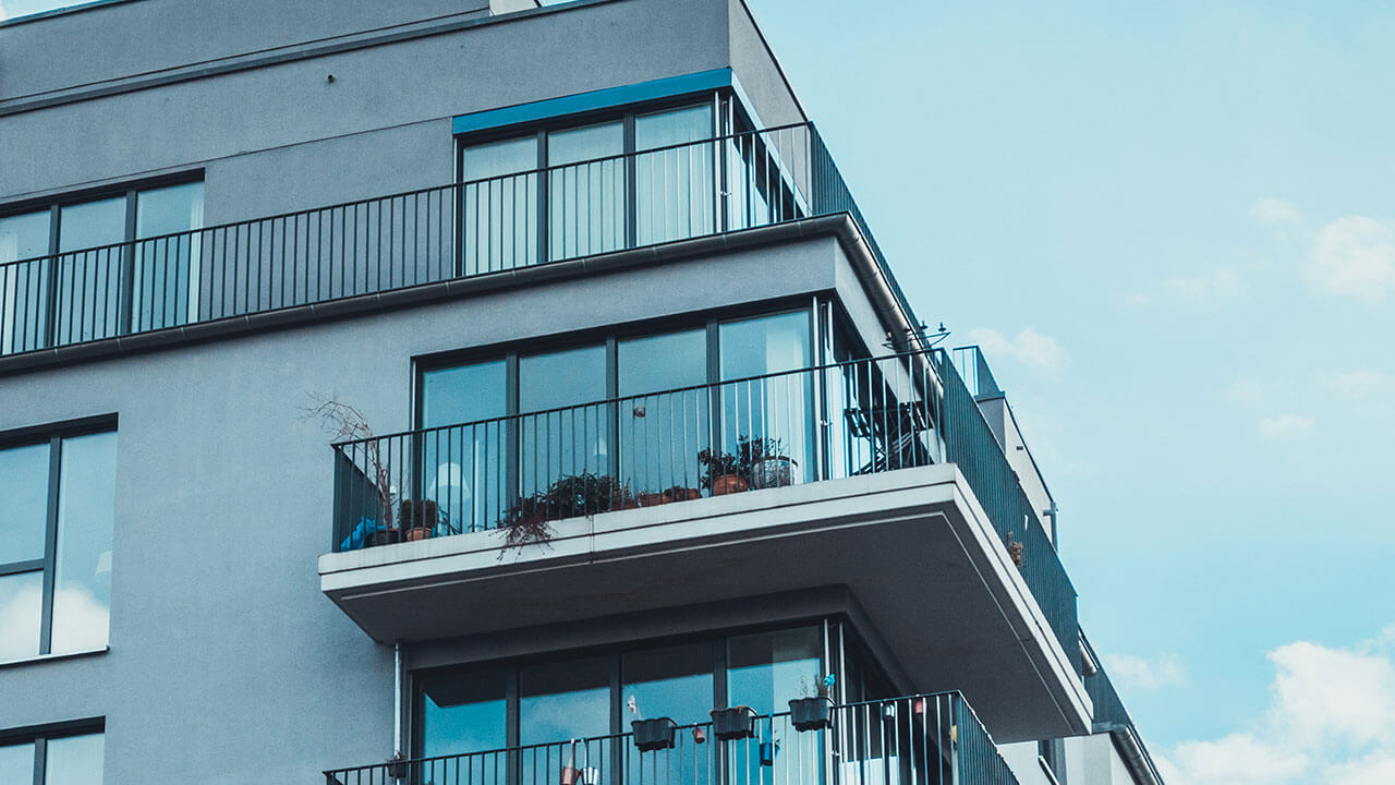 Modern building exterior featuring large glass windows and balconies with potted plants, against a blue sky.