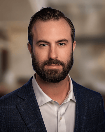 Man with a beard wearing a dark blazer and white shirt, set against a blurred background.