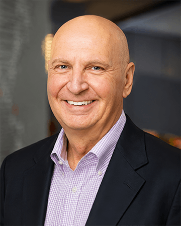 Portrait of a smiling bald man in a black blazer and purple shirt, with a blurred background.