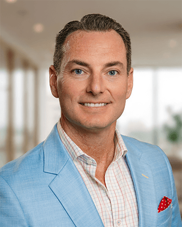 Headshot of a man in a light blue blazer and checkered shirt, smiling in a bright office setting.