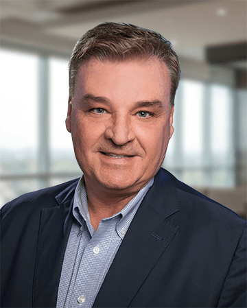 Professional headshot of a man with short hair, wearing a navy blazer over a light blue shirt, with a blurred office background.