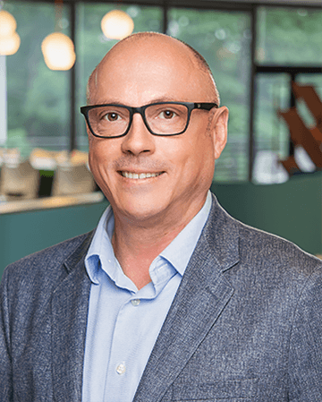 Professional portrait of a man with glasses, smiling, wearing a blazer, in a modern office environment.