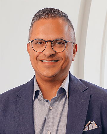 Professional portrait of a man with glasses in a blazer, smiling in a modern office setting.