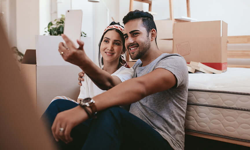 A couple happily takes a selfie while sitting on the floor surrounded by moving boxes.