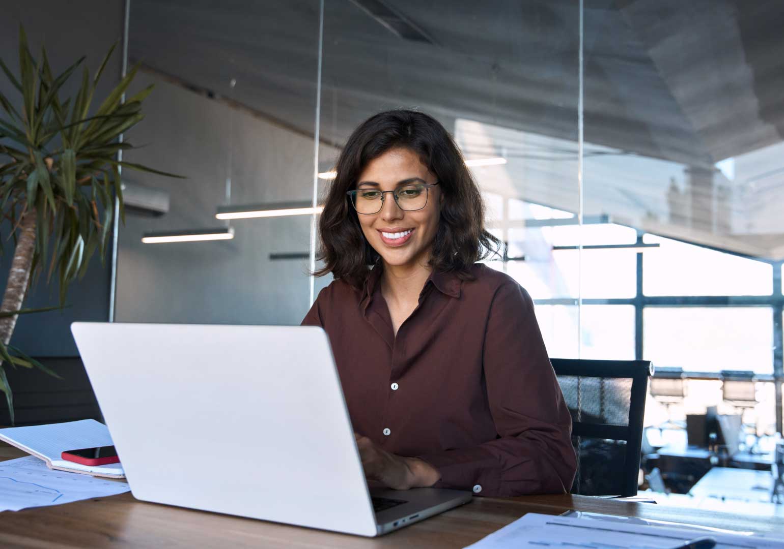 Smiling woman with glasses working on a laptop at a desk in a modern office setting.