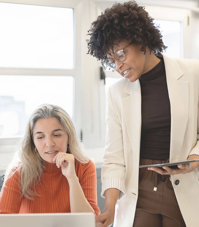 A woman in a white blazer discusses a laptop with another woman in an orange sweater.