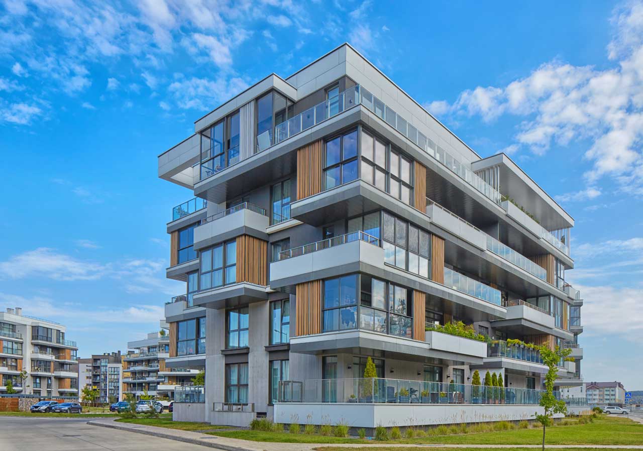 Modern apartment building with large windows and balconies, surrounded by greenery and blue sky.