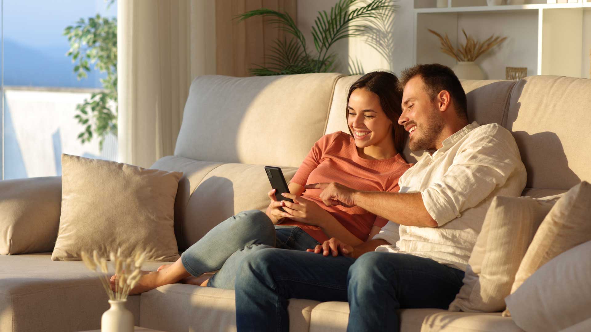 A couple sits together on a couch, smiling while looking at a smartphone, with a bright, sunny interior.