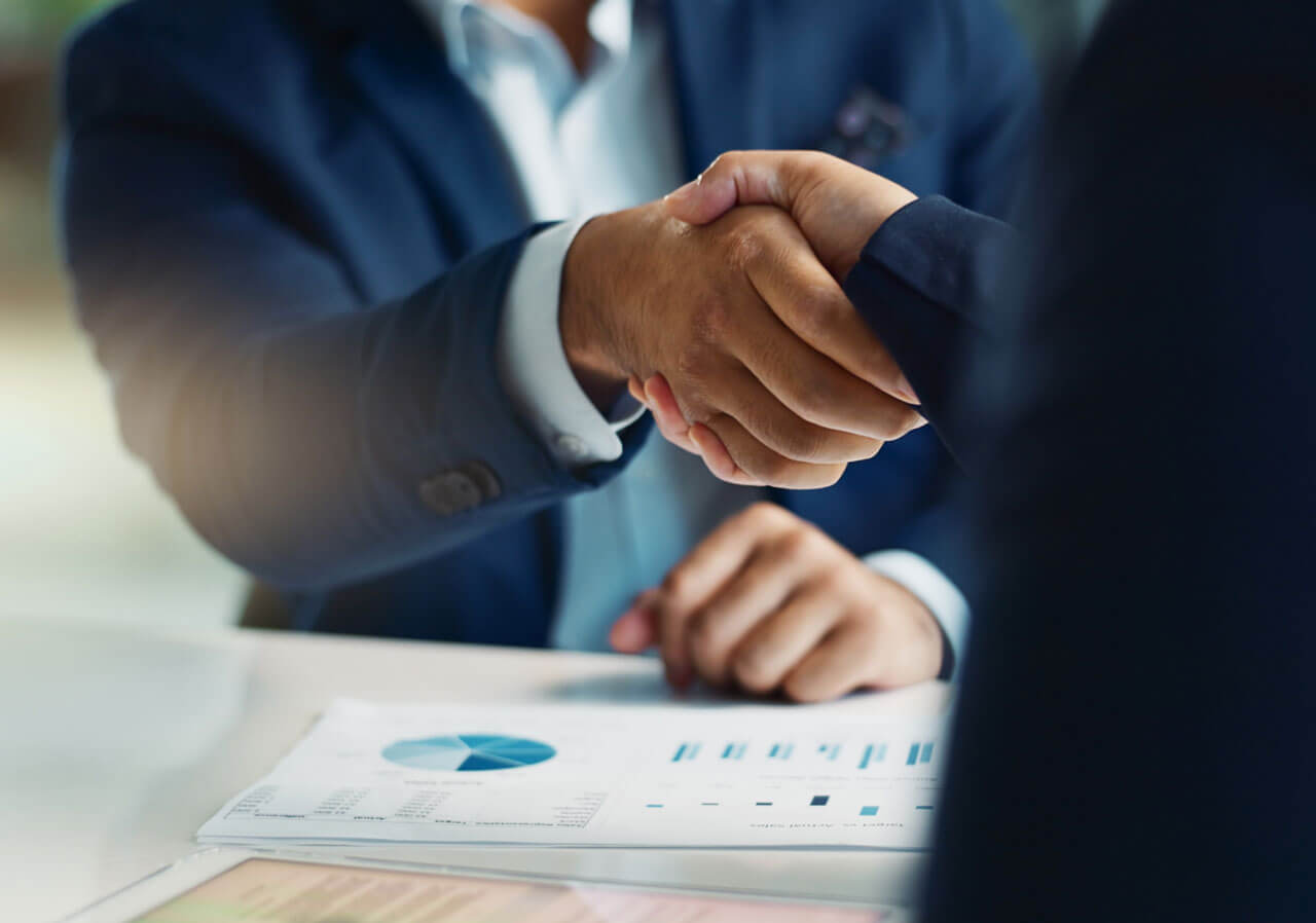 Two business professionals shaking hands over a document with graphs and charts on a table.