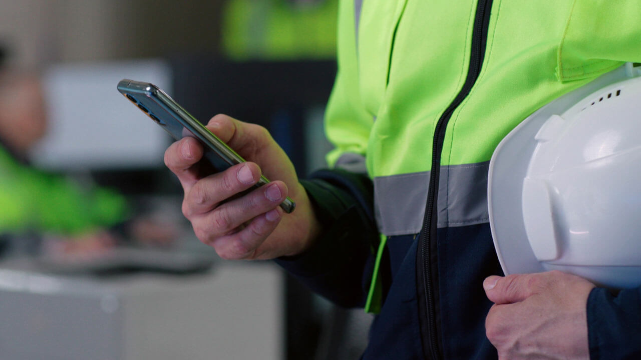 A worker in a high visibility jacket holding a smartphone and a white hard hat in a busy work environment.