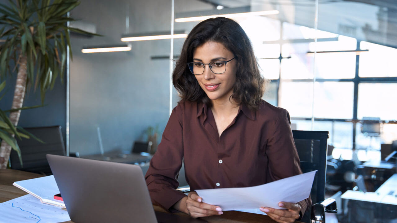 A woman in a brown shirt sits at a desk, reviewing papers while looking at her laptop in a modern office.
