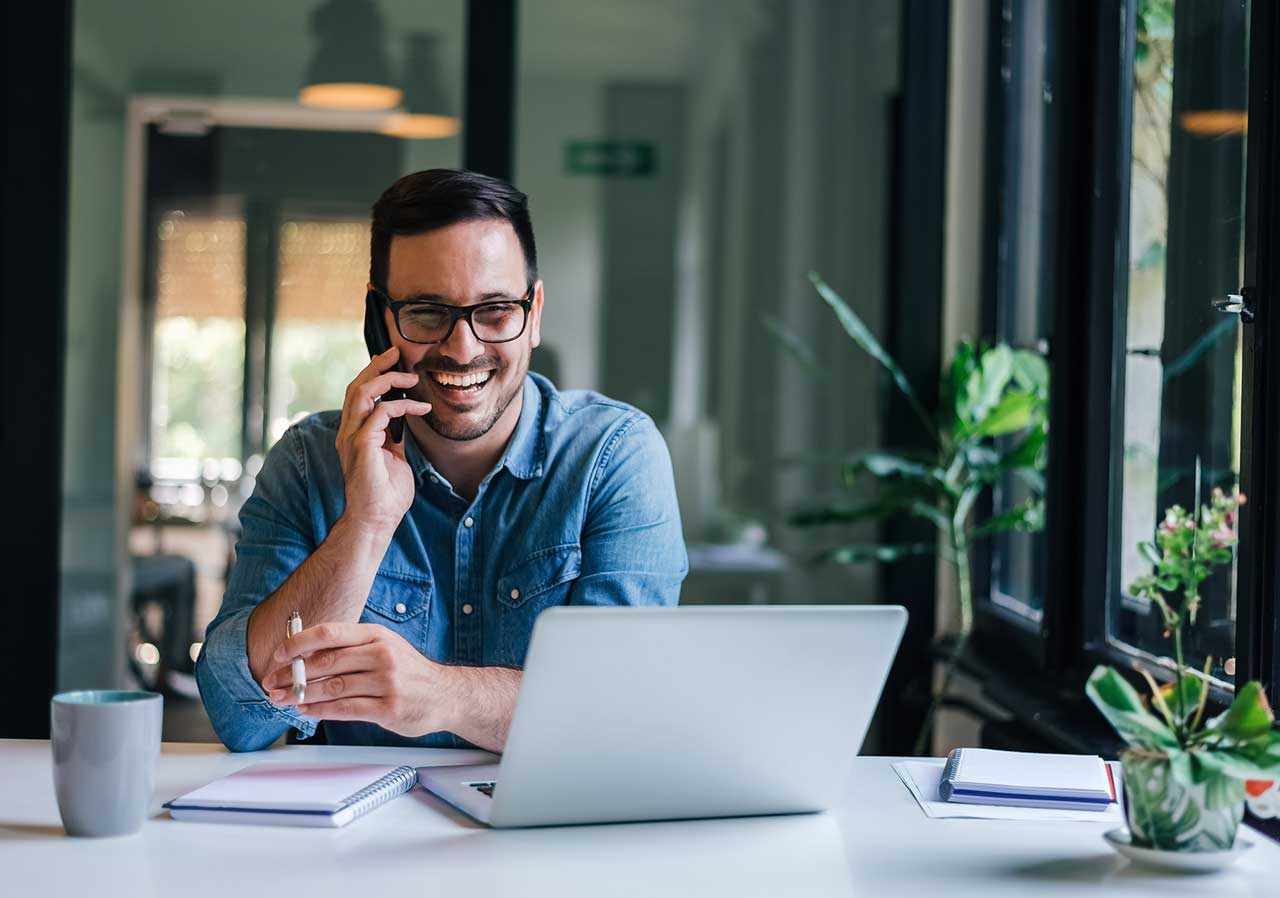 A man in a denim shirt smiles while talking on his phone, working on a laptop at a bright desk with plants.
