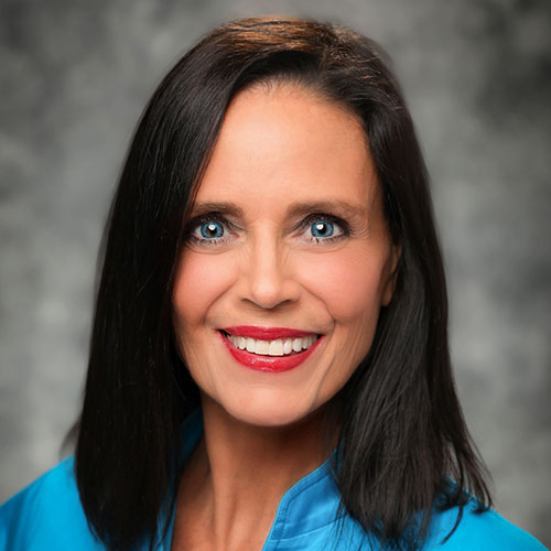 Professional headshot of a woman with long dark hair, wearing a blue blazer, smiling with bright blue eyes.