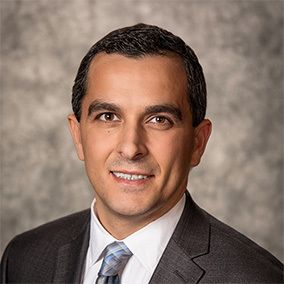 Man in a suit with a tie, smiling against a neutral gray background.