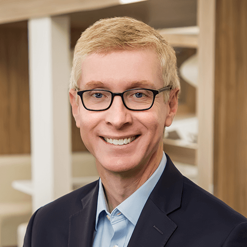 A light-haired man wearing glasses and a dark blazer smiles in an office setting.