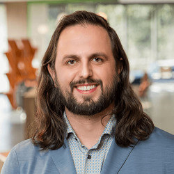 Man with long hair and beard, smiling, wearing a light blue blazer and patterned shirt in a modern indoor setting.