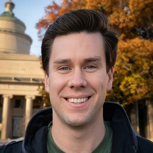 A man with dark hair and a warm smile in front of a building with autumn foliage.